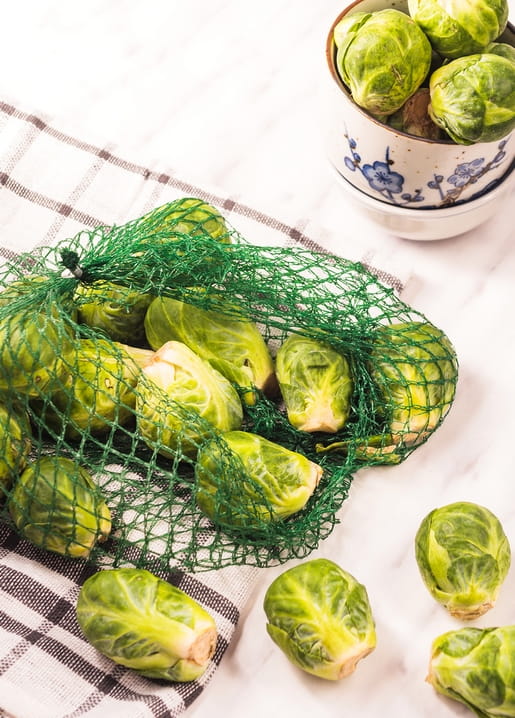 Frischer Rosenkohl im grünen Netz neben einer Porzellanschale auf einem Küchentuch