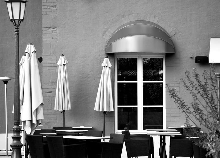 Schwarz-weißes Foto geschlossener Café-Sonnenschirme und leerer Stühle neben einem Bogenfenster mit Vordach