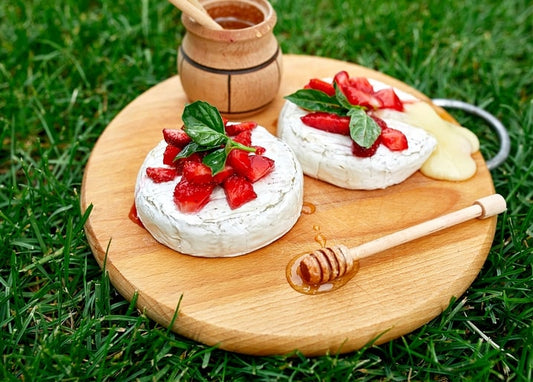 Camembert topped with strawberries and basil, honey dipper on a wooden board in grass