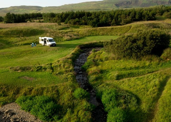 Campervan parked beside a stream in a green valley with distant hills