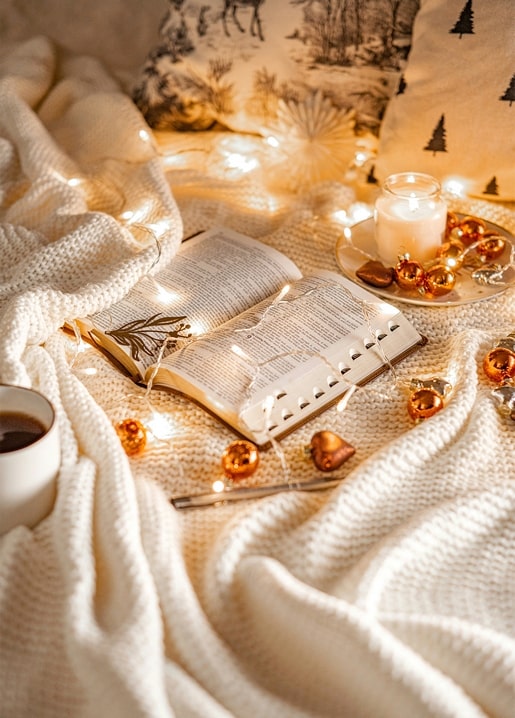 Open book wrapped in fairy lights beside a lit candle and copper baubles on a cream knit blanket