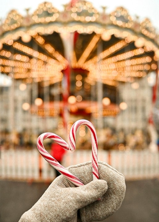 Two candy canes shaped into a heart, held in a mitten with a lit carousel blurred behind