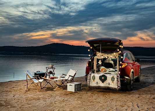 Red car with open trunk decorated with fairy lights beside a beach picnic at sunset