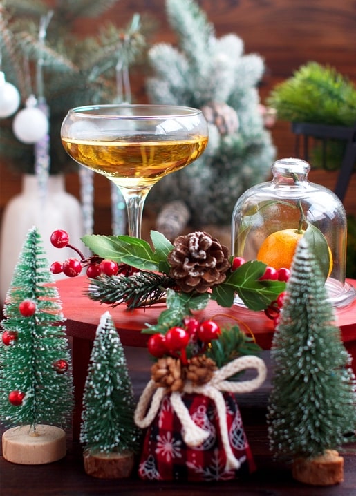 Glass of sparkling wine, mini Christmas trees, pinecones and a tangerine under a glass cloche