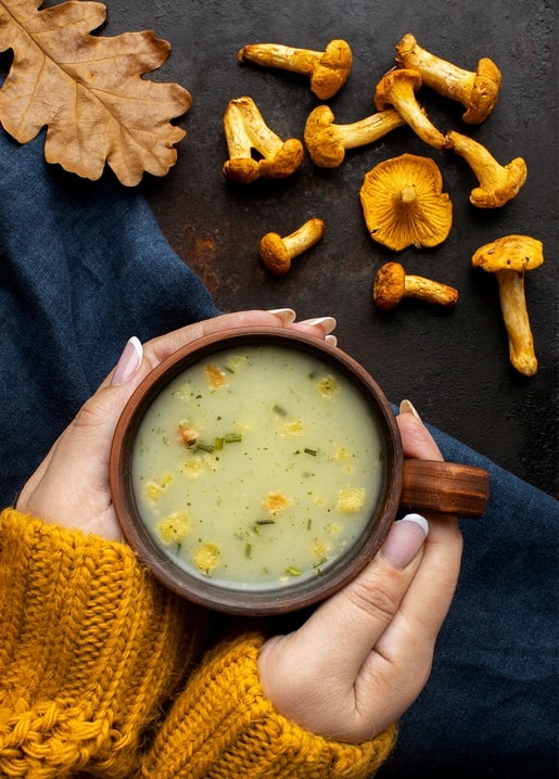 Hände halten einen Holzbecher mit Suppe neben Pfifferlingen und einem Eichenblatt