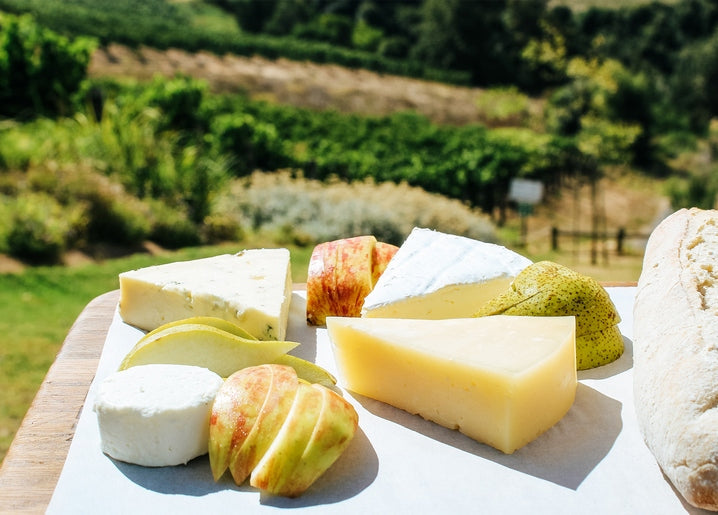 Wooden board with blue cheese, brie, pears, apples, and bread in front of sunny vineyard hills