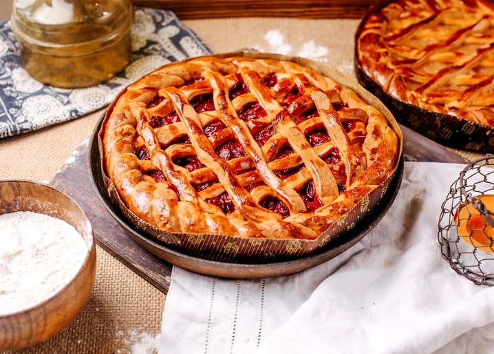 A golden cherry pie with a lattice crust on a rustic table, flour and a second pie nearby.