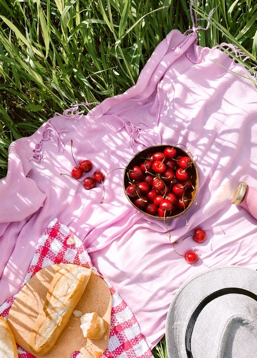 Kirschen, Brot und ein Strohhut auf einer rosa Picknickdecke im Gras