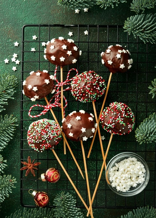 Chocolate cake pops with star sprinkles on a cooling rack among pine branches and ornaments