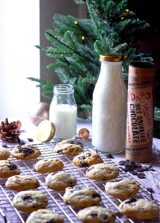 Chocolate chip cookies cooling on a rack beside milk and hot chocolate near a lit pine branch