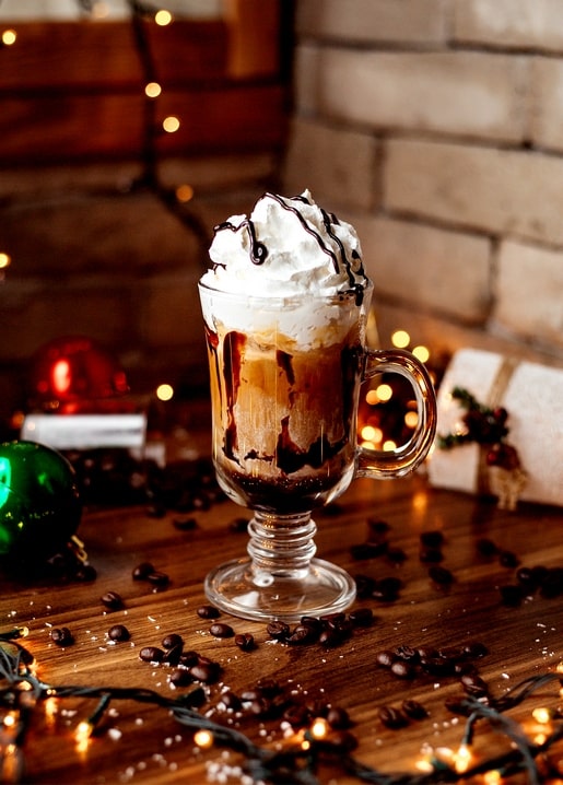 Hot cocoa with whipped cream and chocolate drizzle beside Christmas ornaments and coffee beans on a wooden table