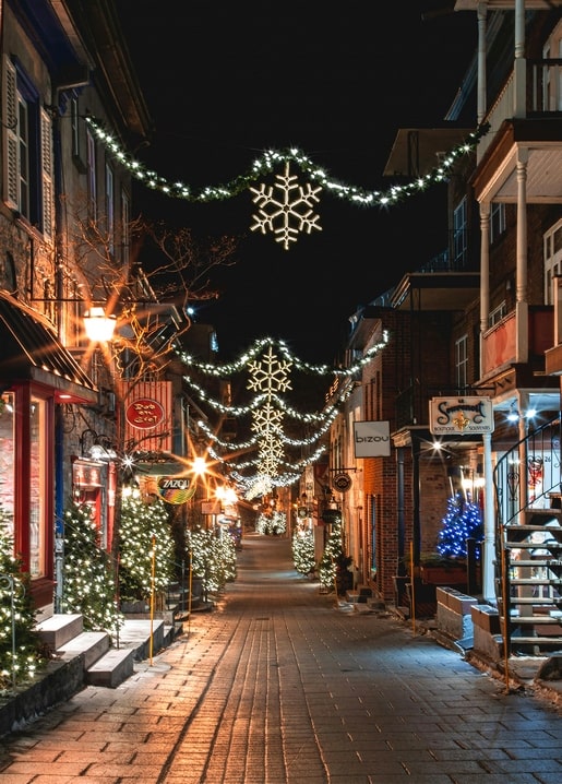 String lights and lit snowflake ornaments over a cobblestone Christmas market street at night