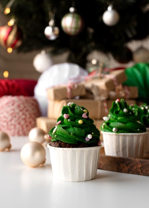 Two chocolate cupcakes with green Christmas tree frosting and sprinkles near ornaments and gifts