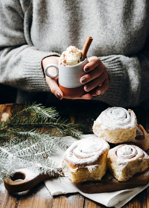 Hands in a grey sweater holding cocoa beside cinnamon rolls and pine