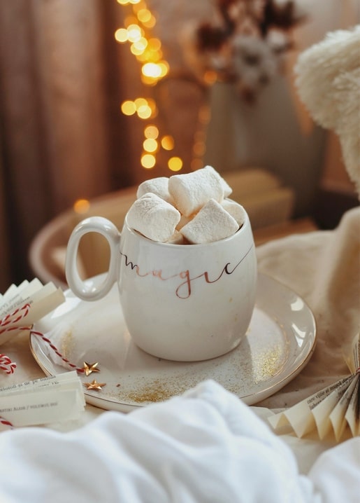 White mug lettered 'magic' filled with marshmallows beside fairy lights