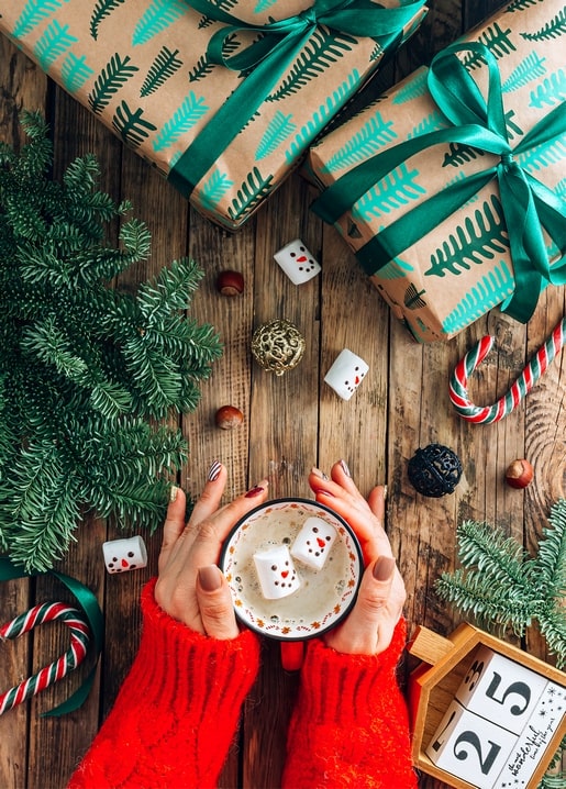 Hands in a red sweater holding cocoa with snowman marshmallows beside wrapped Christmas gifts
