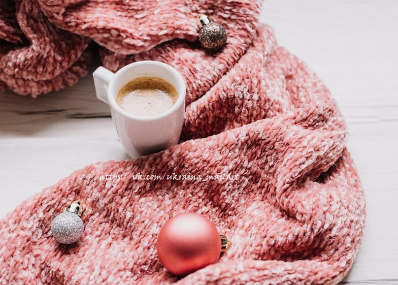 Tasse Kaffee mit silberner und roter Christbaumkugel auf einer rosa Strickdecke