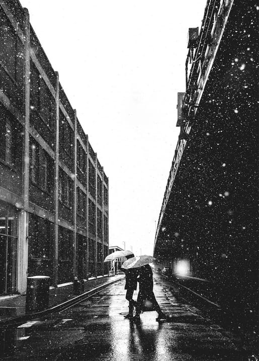 Couple sharing an umbrella crossing a snowy city street at night beneath an elevated trestle