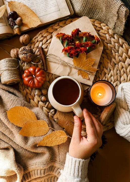 Hand hält eine Teetasse neben Kerze, Ringelblumen und kleinem Kürbis
