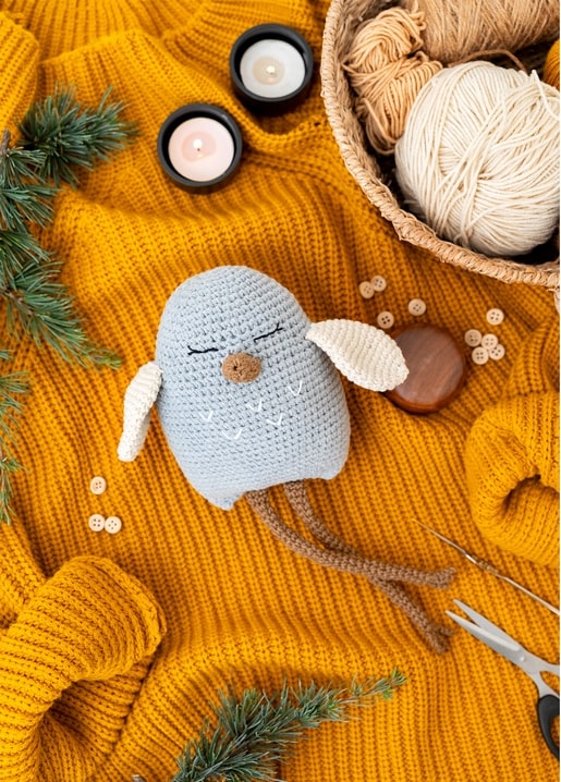 Gehäkelte Vogelfigur auf senffarbener Strickdecke mit Kerzen, Garn und Kiefernzweigen