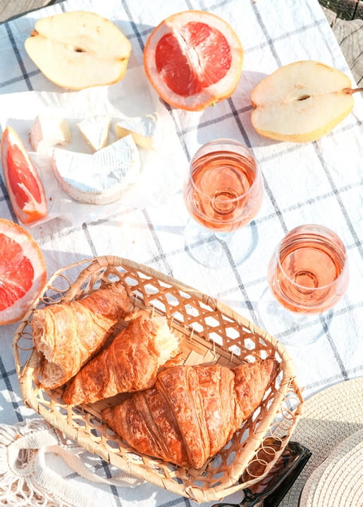 Karierte Picknickdecke mit Croissants, Brie, halbierten Birnen, Grapefruit und Gläsern Rosé