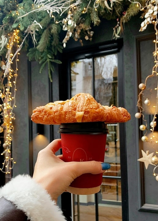 Hand holding a croissant balanced on a red coffee cup outside a decorated café