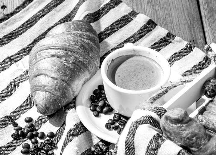 Schwarz-weißes Foto von Croissant, Espressotasse und Kaffeebohnen auf gestreiftem Tuch