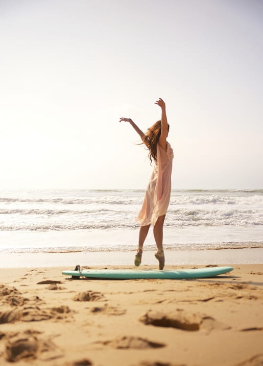 Frau im wehenden Kleid tanzt am Strand neben einem türkisfarbenen Surfbrett