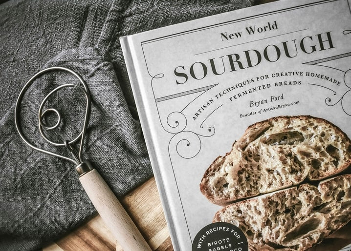 Teigbesen aus Draht neben einem Sauerteig-Kochbuch und aufgeschnittenem Brot