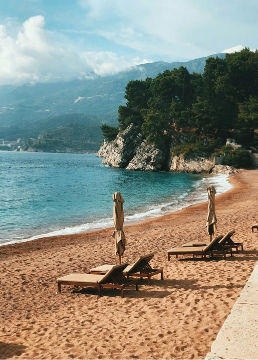 Leerer Strand mit Holzliegen und geschlossenen Sonnenschirmen vor türkisfarbener Bucht
