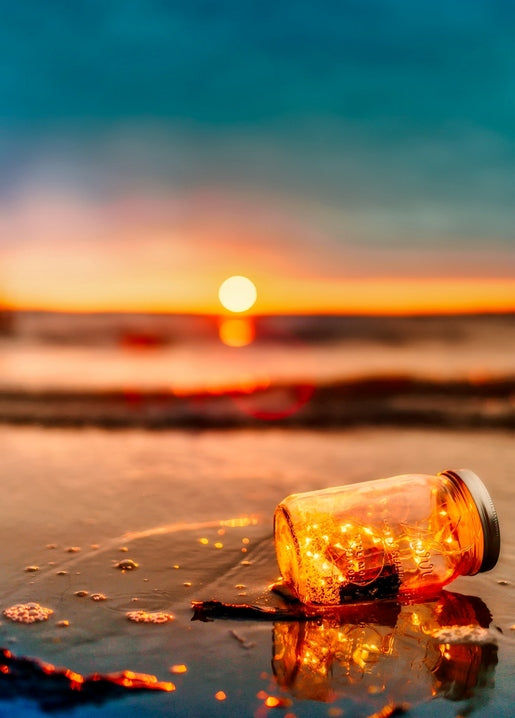 Glass jar filled with warm fairy lights sitting on wet sand at sunset, with the sun reflected on the sea.