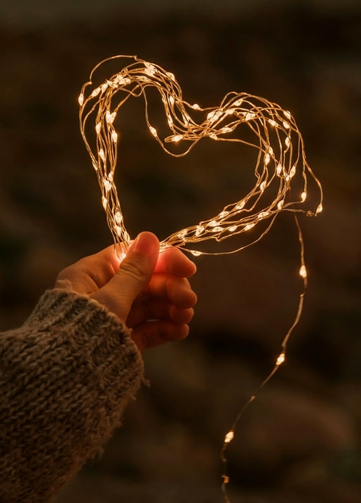Hand in a knit sleeve holding a heart shaped from warm fairy lights against a dark background