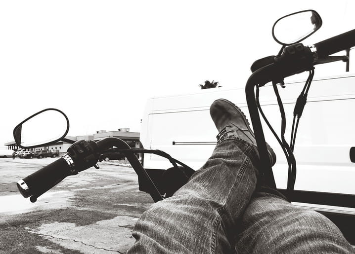 Black-and-white photo of feet resting on bicycle handlebars beside a parked van