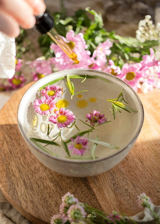 Pink daisies and rosemary sprigs floating in a bowl of water with a dropper adding oil