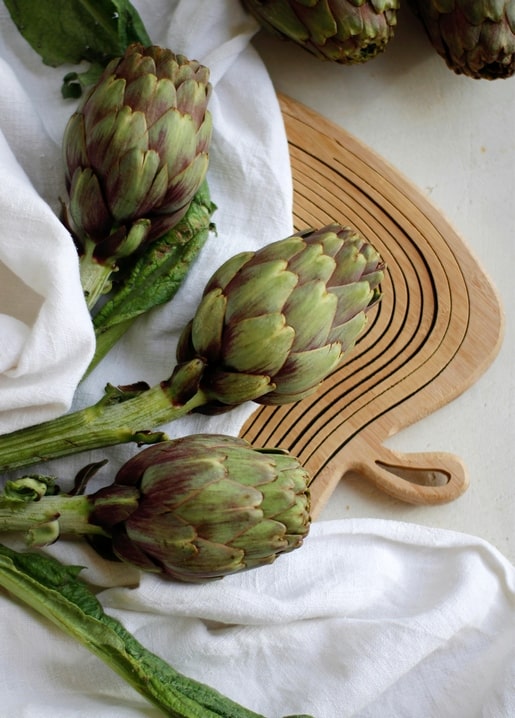 Three fresh artichokes with stems and leaves on a rustic wooden board wrapped in white linen