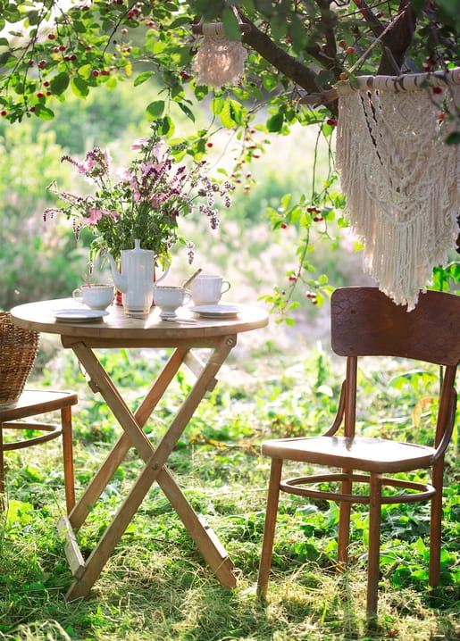Gartentisch mit Teegeschirr, Wildblumen im Krug und Makramee, das vom Baum herabhängt