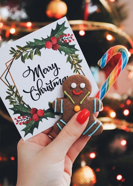Gingerbread man cookie and candy cane beside a hand-held Merry Christmas card with holly and tree lights