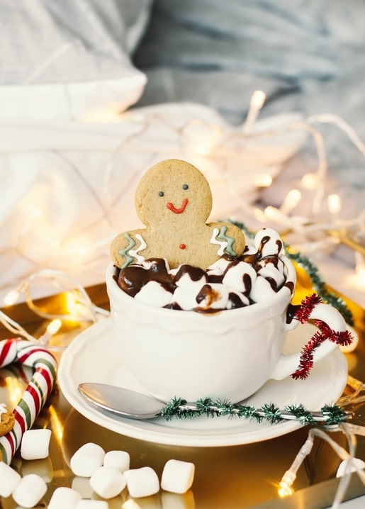 Gingerbread man cookie in a mug of hot cocoa with marshmallows and fairy lights