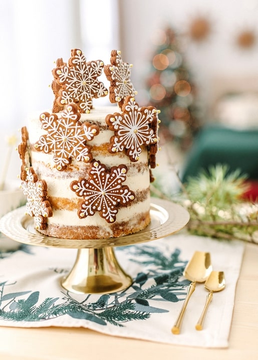Naked layer cake topped with iced gingerbread snowflakes on a gold stand with pine and Christmas lights