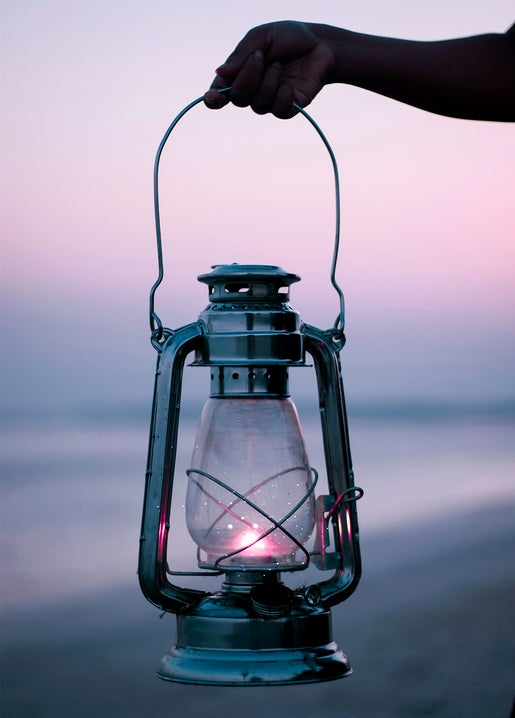 A hand holds a glowing oil lantern against a pink-violet dusk sky over the sea.