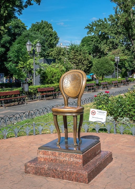 Gilded bronze chair sculpture on a marble pedestal in a sunlit Odesa park.