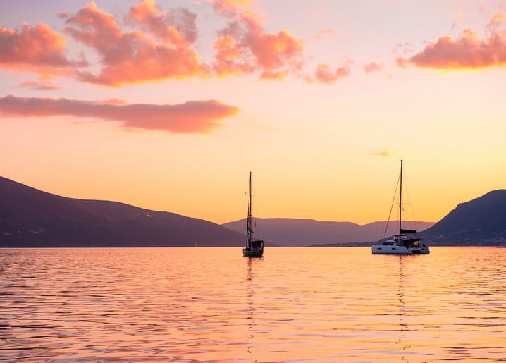 Zwei Segelboote auf einer ruhigen Bucht bei Sonnenuntergang, Berge als Silhouette vor rosa-goldenem Himmel