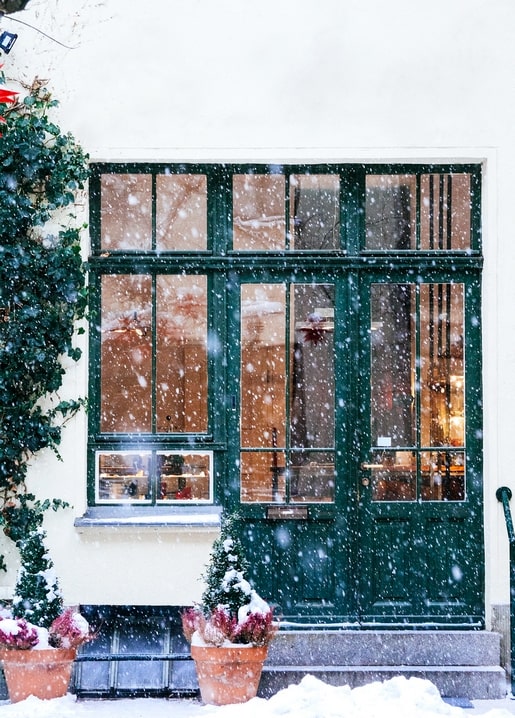 Snow falling past a dark green door and windows with ivy and potted firs