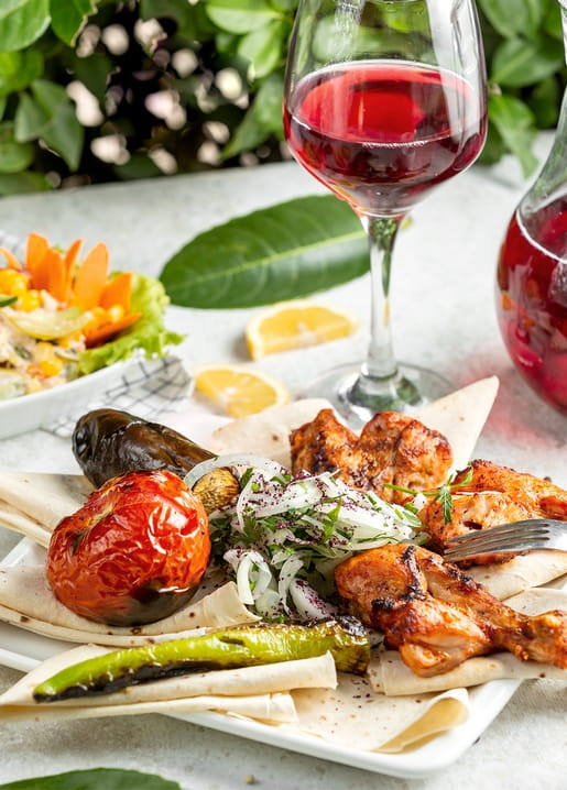 Grilled chicken skewers, roasted vegetables, pita and a glass of red wine on a table