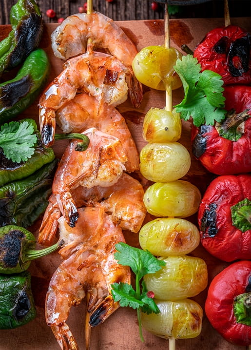 Draufsicht auf gegrillte Garnelen, Kirschtomatenspieße, geröstete Paprika und Koriander auf einem Holzbrett