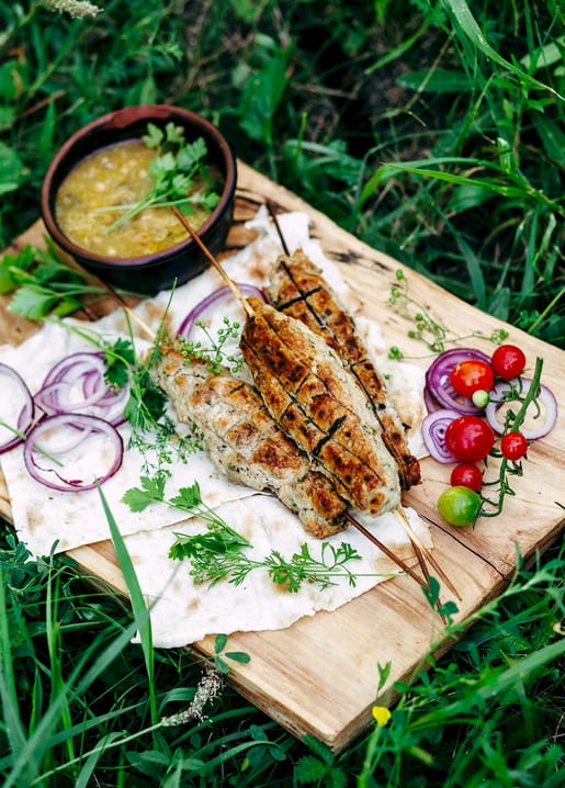 Gegrillte Fleischspieße mit Kräutersoße, roten Zwiebeln und Tomaten auf einem Holzbrett im Gras