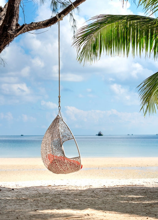 Empty rattan hanging chair swinging beneath palm fronds on a white-sand beach, turquoise sea beyond.