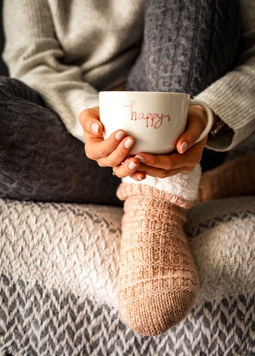 Hände halten eine weiße Tasse mit Aufschrift „happy“ neben Strickstrümpfen auf einer Decke