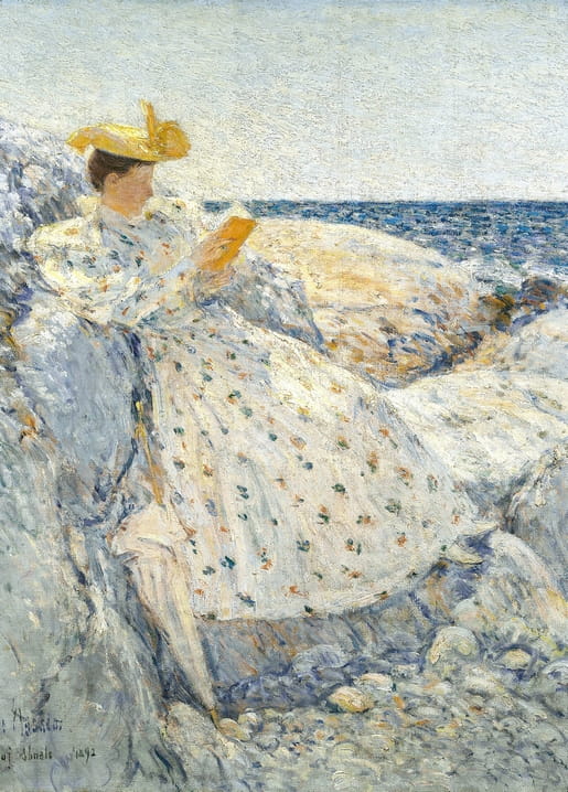Woman in a dotted white dress and yellow hat reading a book on sunlit rocks by the sea