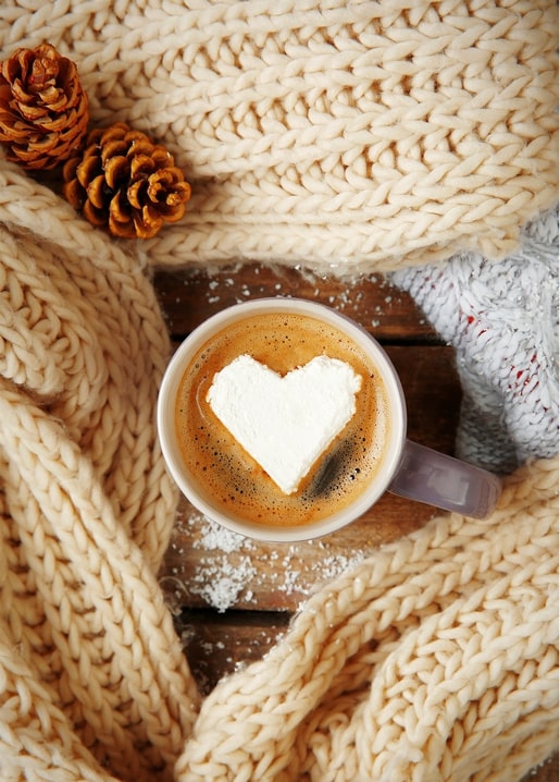Latte with heart-shaped foam wrapped in a chunky cream knit beside pinecones